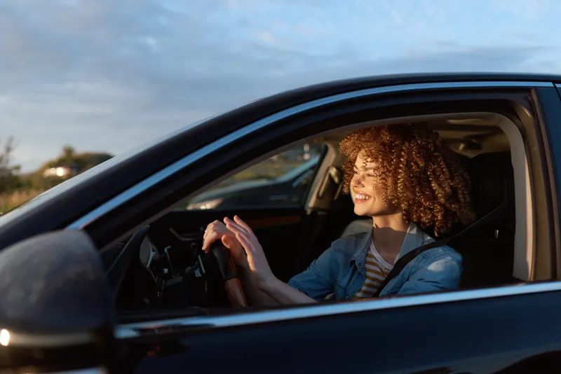 Young woman with curly hair driving a car in daylight, smiling at the wheel. Casual lifestyle scene conveying freedom, daily routine, and self expression.
