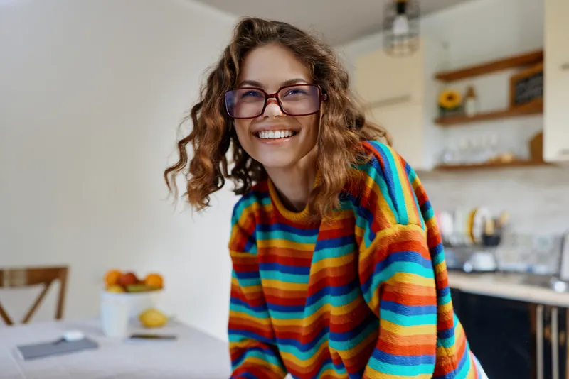 A woman with curly hair wearing a colorful striped sweater smiles warmly in a bright modern kitchen, leaning toward the camera as if inviting a friendly chat and sharing a light moment.