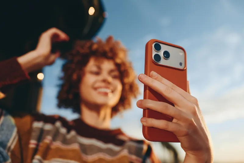 Woman lifestyle selfie smartphone curly smiling hand outdoors portrait technology, woman holds smartphone for selfie, curly hair and striped sweater in soft blur, friend waving behind, bright