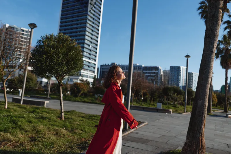 A woman in a red coat walks through a sunny urban park, palm trees and modern buildings in the background. A candid moment of daily life in the city.
