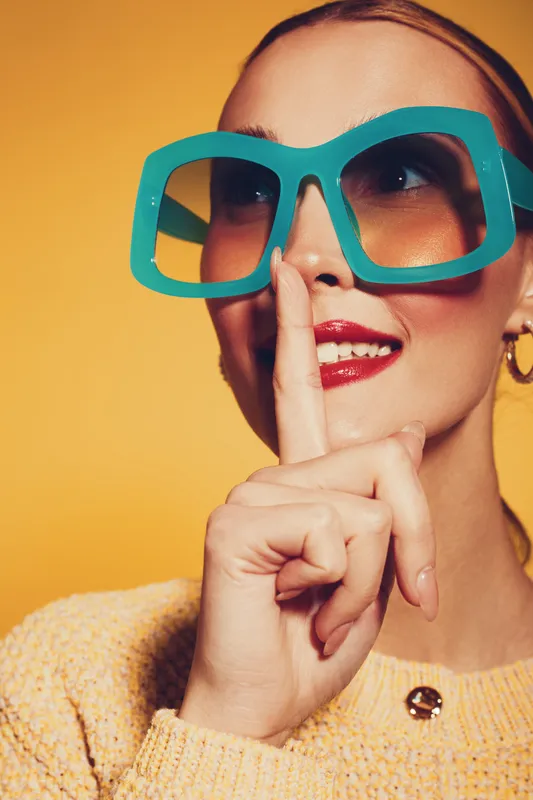 Young woman with large turquoise glasses holds finger to lips in shushing gesture. She wears a yellow sweater and gold hoop earrings against a bright yellow background. Concept of secrecy.