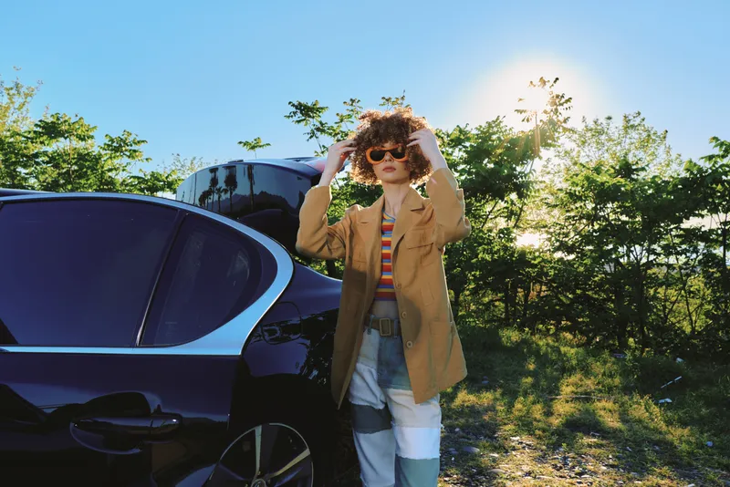 A stylish woman in a tan coat and striped shirt stands beside a dark car, sunglasses on, under bright sun. Retro film color mood enhances a casual lifestyle and outdoor moment.