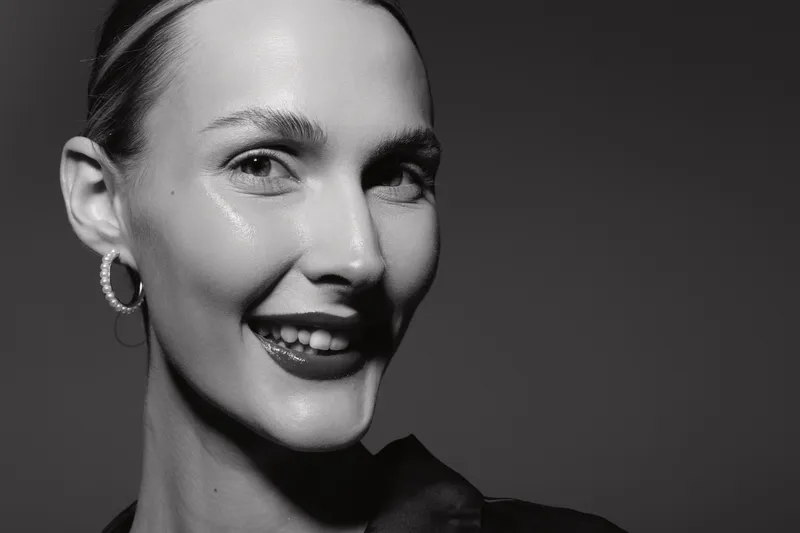 Close-up portrait of a smiling Caucasian woman with light skin, wearing hoop earrings, against dark background. Studio lighting highlights facial features.