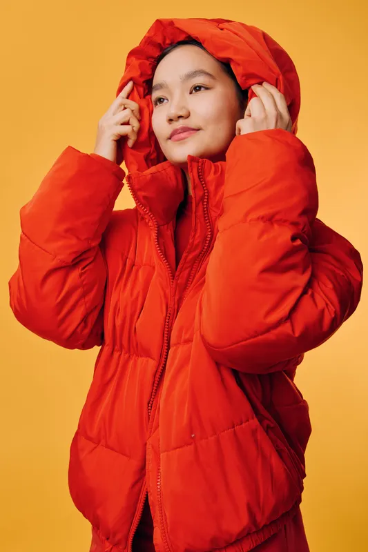 Young woman wearing red puffer jacket with hood on yellow background. Portrait, casual style, winter clothing, cold weather outfit, confident expression, fashion apparel.