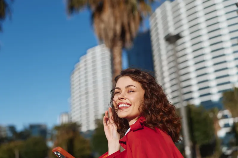 Young woman with curly hair smiles happily while holding a smartphone outdoors in an urban setting on a sunny day.
