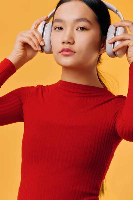 Young woman with long hair wearing large headphones and red sweater posing against orange background for music, technology, and lifestyle concept portrait.