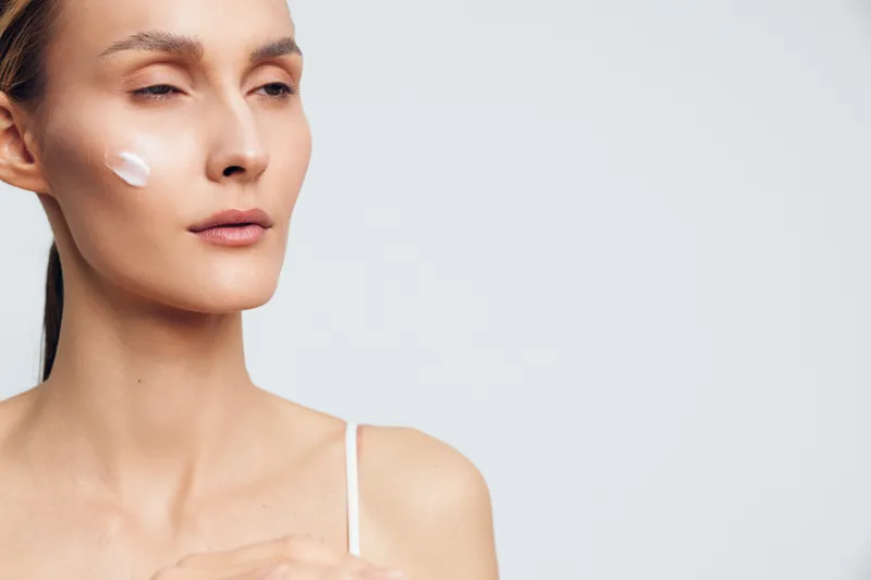 Natural beauty portrait of a woman with skincare cream on cheek, minimal makeup, soft lighting, studio background, showcasing a simple daily skin care routine and calm mood