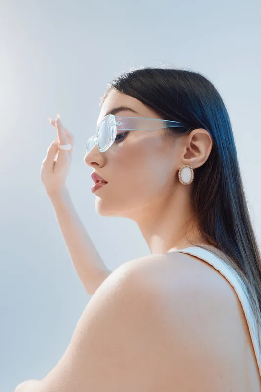 Profile of a young woman with long dark hair wearing transparent glasses and elegant earrings. She poses gracefully against a soft light background. Concept of beauty fashion.