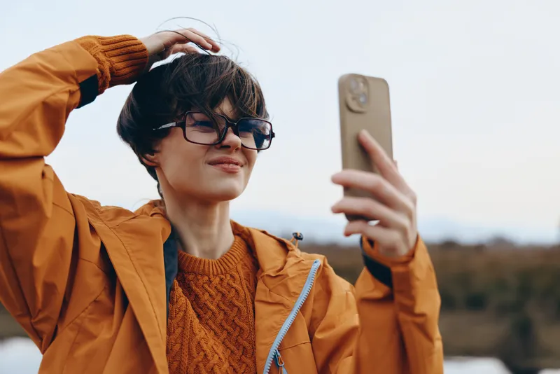 Woman lifestyle outdoor selfie, woman adjusts hair with one hand and wears sunglasses, orange jacket and knit sweater, holding smartphone for portrait, autumn background blur, casual confident mood