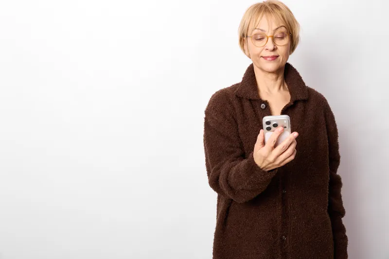 Portrait of an elderly woman with glasses, holding a smartphone, calm expression, using texting for communication, minimalist studio background, technology concept
