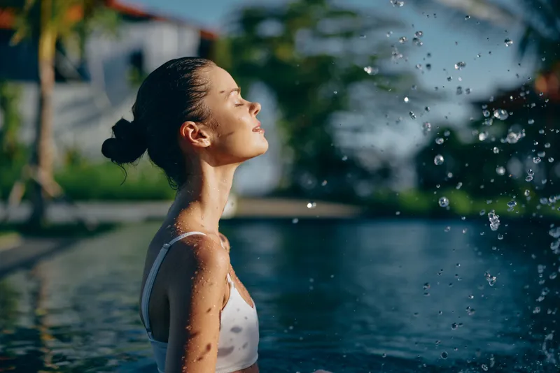 Cheerful woman outdoors by a pool, eyes closed, savoring sunlight, droplets in the air, relaxed posture, warm breeze, lush backdrop, vibrant colors, lifestyle moment, serene and joyful