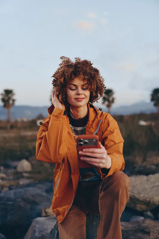Woman lifestyle outdoors portrait, curly hair, orange jacket, holding phone, smiling in relaxed stance on rocks at sunset, golden light, calm mood with distant hills and clear sky