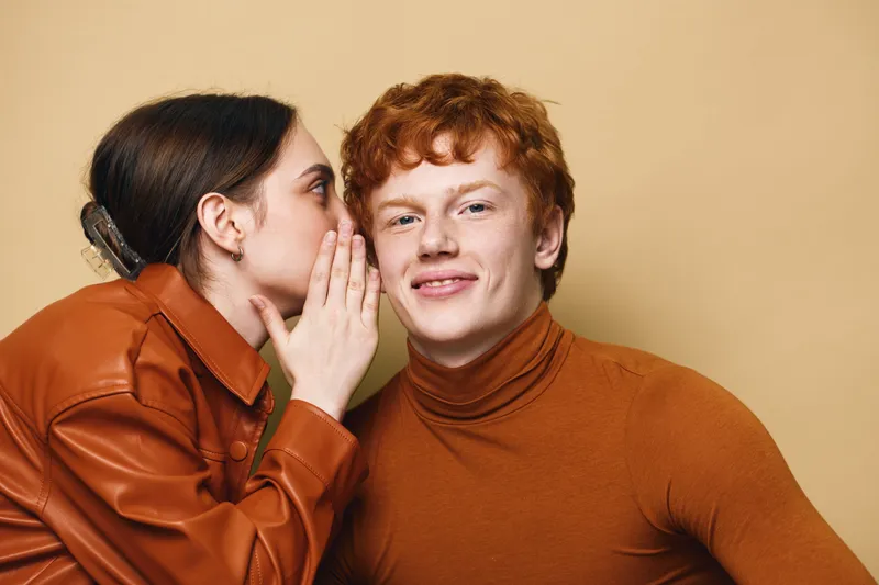 Young woman whispering secret to smiling redhead man with casual clothes, friendship, communication and closeup portrait on beige background with emotions and sharing news.