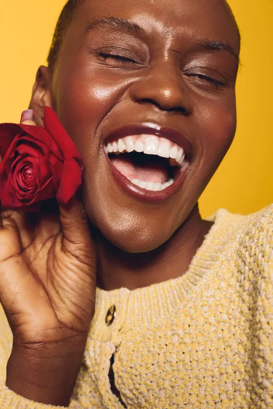 Happy dark skinned African woman laughing with closed eyes holding red rose near face. Close up on yellow background. Concept of joy, beauty, natural skin, and floral happiness.