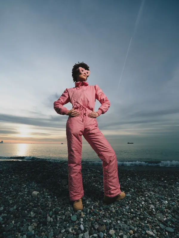 Pink jumpsuit worn by a person standing confidently on a pebbled beach at sunset, embracing outdoor scenery, calm sea, and a dramatic sky above the horizon.
