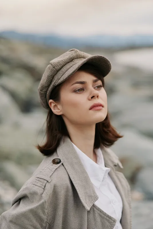 Young woman in vintage coat and cap looks thoughtfully into distance outdoors. Natural makeup and soft light highlight her contemplative expression and autumn style fashion concept.