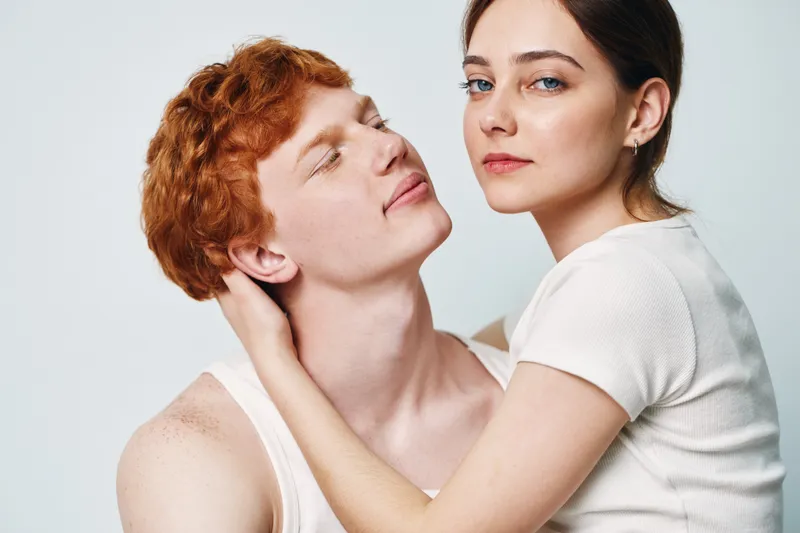 Young couple embracing in closeup with woman holding redhead man, both looking calm and connected, casual clothing on light background for relationship and intimacy.