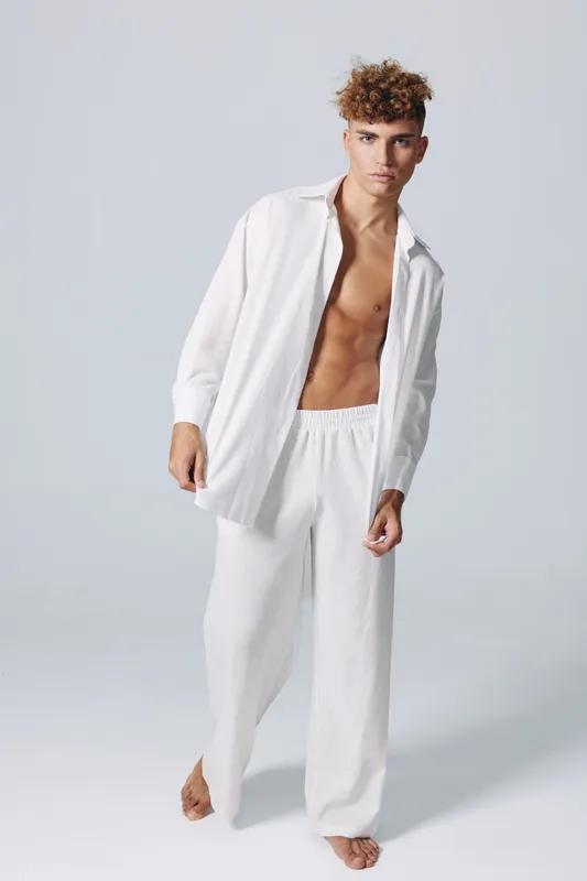Young man with curly hair wearing loose white pants and an open white shirt standing barefoot on a plain background with a confident and relaxed pose.