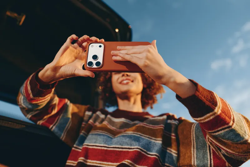 Woman lifestyle smartphone selfie, hands holding phone for closeup portrait, curly haired woman smiling in soft focus behind, outdoor daylight, striped sweater, technology moment