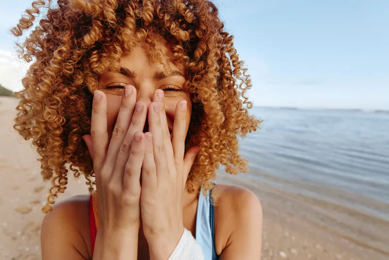 Joyful woman with curly hair on a sunny beach covers her face with hands, revealing laughter and surprise in a candid seaside portrait on a calm shore