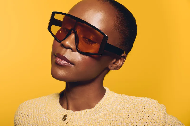 Confident African woman with dark skin wearing large brown sunglasses and a beige knitted sweater poses against yellow background. Concept of fashion, style, and modern eyewear.