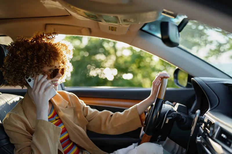 Woman driver sits in a vintage car talking on the phone, casual lifestyle scene with retro film color grading, sunlight through window, stylish striped shirt and jacket