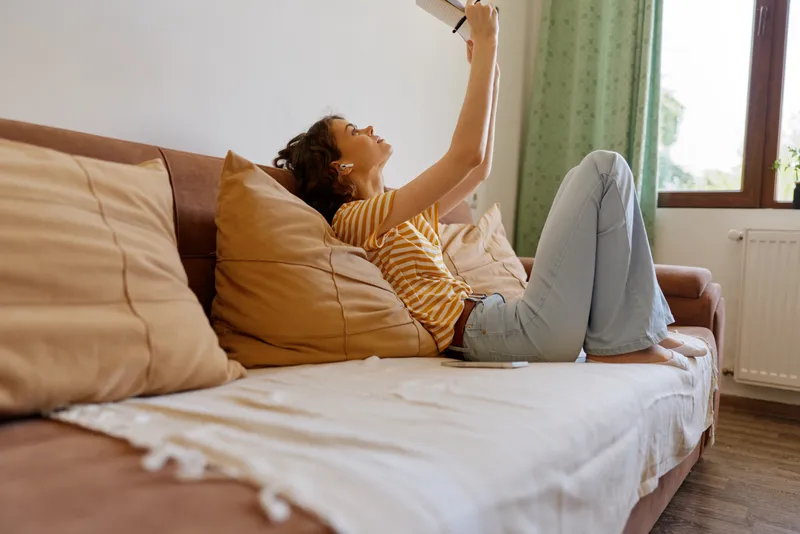 Relaxed woman lounges on a plush sofa in a bright living room, stretching her arms to reach a remote control with a content smile, casual jeans, striped shirt, warm natural light