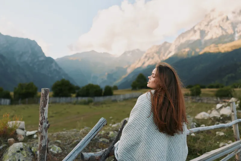 A young woman in a knit cardigan gazes toward distant mountains from a weathered wooden fence.