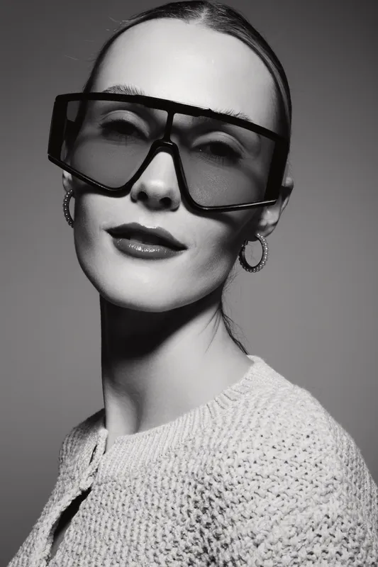 Caucasian woman with fair skin and slicked back hair wears oversized sunglasses and hoop earrings. Studio portrait in black and white lighting.