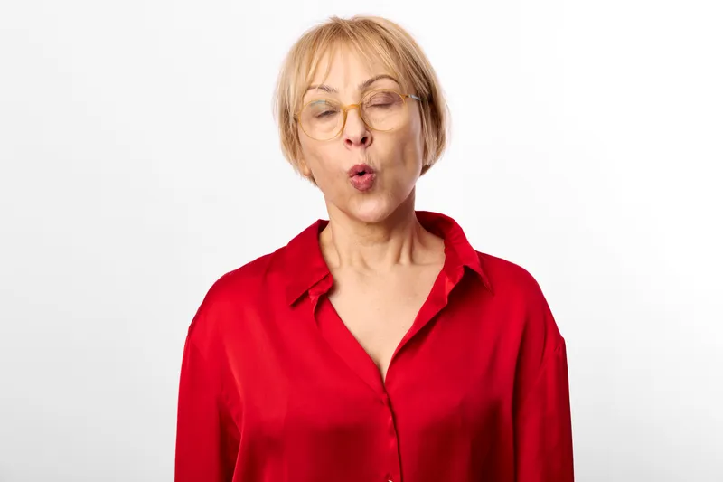 Woman senior surprised face with glasses, wearing red shirt, standing in white background portrait, close up emotion, looking at camera with open mouth directly