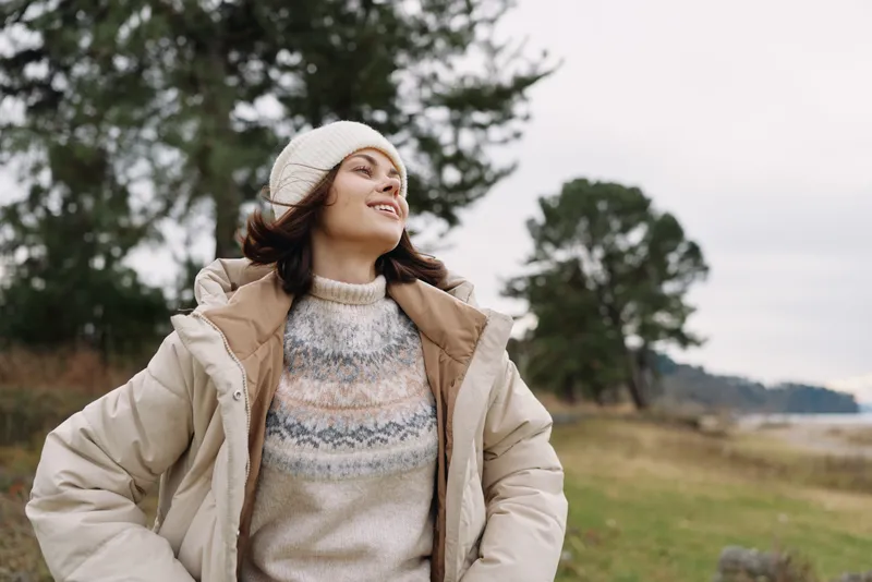 Woman outdoors wearing a warm beige coat, knitted sweater and beret, enjoying fresh air in an open landscape with trees, conveying casual fashion and a hopeful mood.