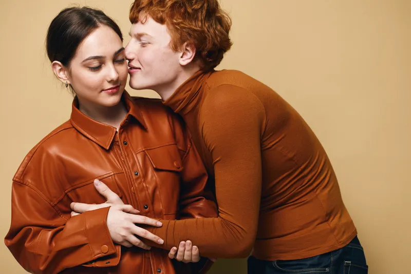 Couple with affection indoors, woman wearing leather jacket and man in brown turtleneck smiling with close embrace for love and intimacy.