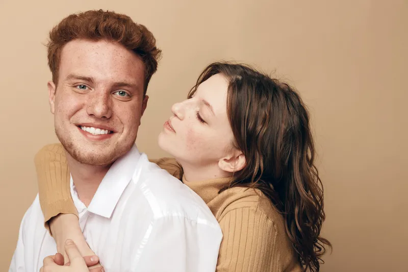 Smiling young man in white shirt embraces woman in brown sweater who looks at him affectionately. Warm beige background creates cozy atmosphere of love and close relationship.