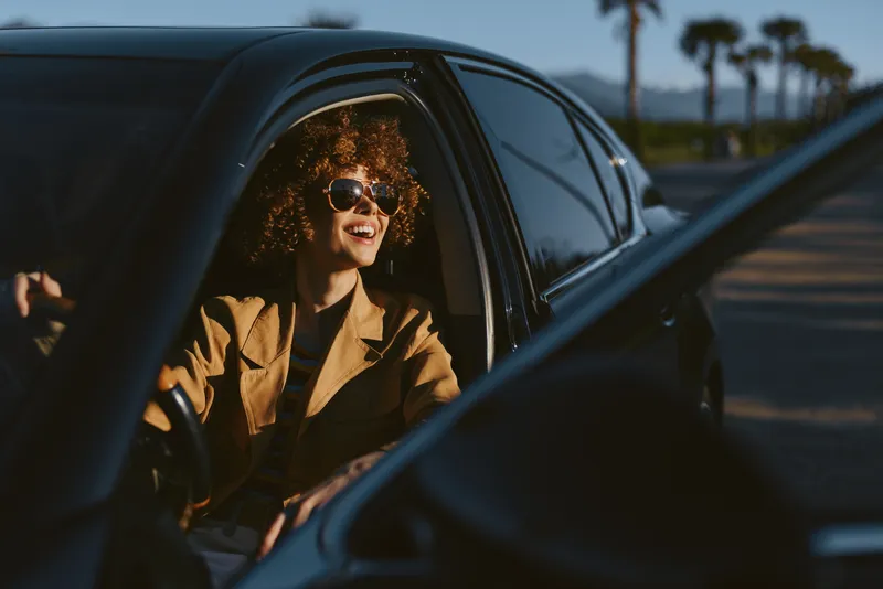 Woman enjoys candid drive, lifestyle scene in retro film color, smiling behind sunglasses inside a car, warm natural light, casual chic mood on the open road, carefree vibe.