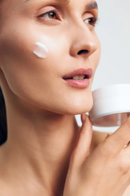 A close-up of a young woman with smooth skin applying white cream on her cheek while holding a cosmetic jar, symbolizing skincare and beauty routine for healthy complexion.