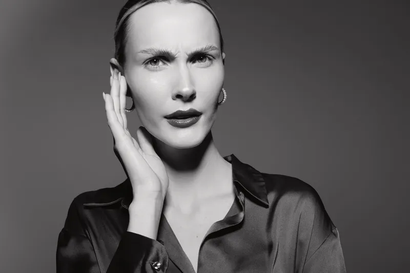 White woman with pale skin and slicked back hair touches face with hand, looking confused or concerned in black silk shirt. Studio portrait in black and white lighting.