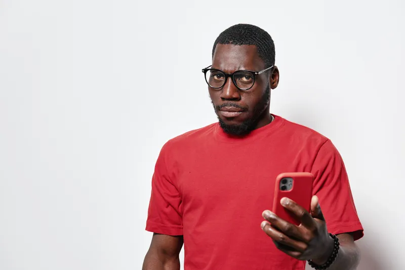African American man with dark skin wearing glasses and red t shirt holding smartphone looking serious on white background. Male adult with beard and short hair using mobile device.