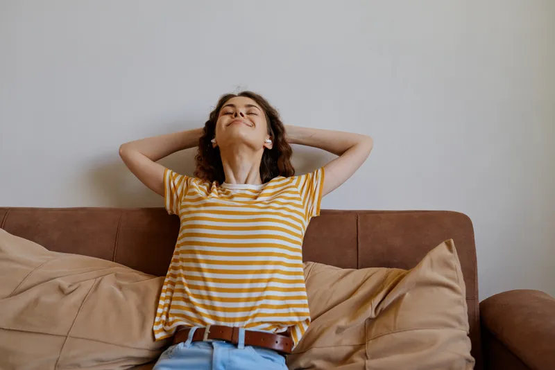 Relaxed woman at home lounging on a sofa in a striped yellow shirt, casual pose with hands behind head, cozy lifestyle scene, warm natural light, peaceful mood and everyday comfort