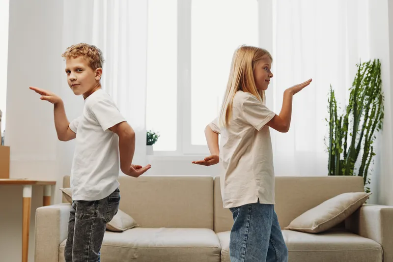 Two children stand back to back indoors, playfully posing with hands raised, casual clothes, bright living room with sofa and plants behind.