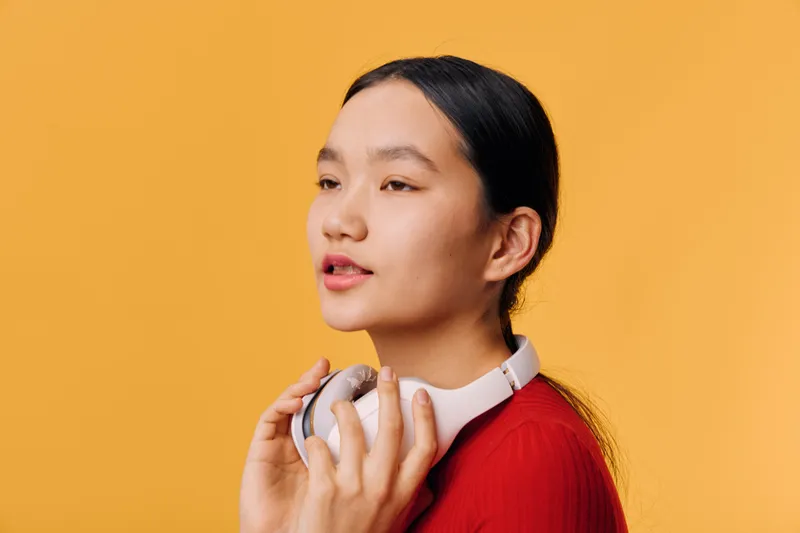 Young woman with long black hair holding white headphones, looking to the side with closed mouth and relaxed expression, wearing red top against yellow background.