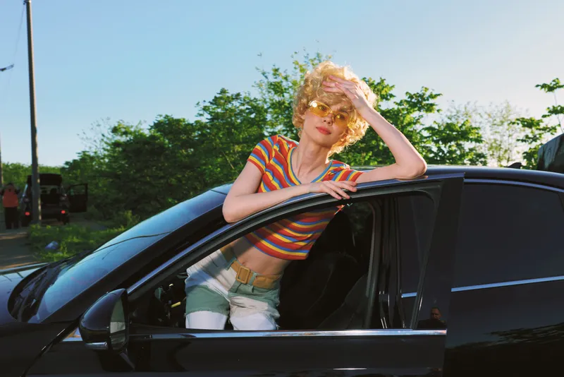 A young woman leans out of a black car window on a sunny afternoon, wearing a striped crop top and shorts, capturing a carefree street style with retro film color vibes.