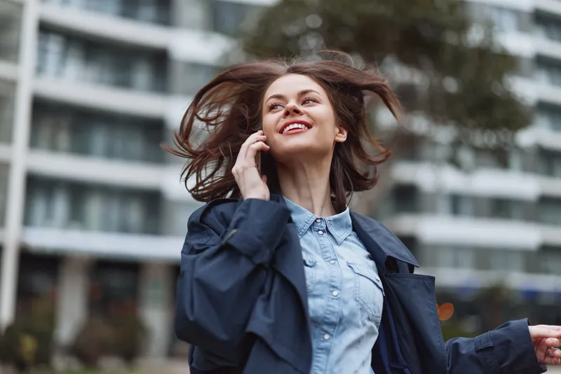 A happy young woman wearing a blue jacket talks on a smartphone outdoors with hair blowing in the wind, smiling against an urban background. Concept for communication and joy.