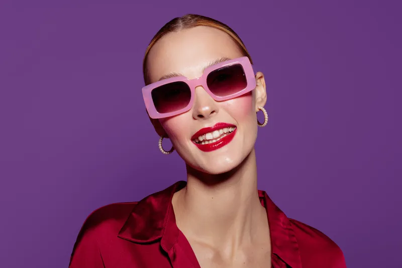 Smiling woman with pink sunglasses and red lipstick poses against purple background. She wears red satin blouse and gold hoop earrings. Concept of fashion, beauty, and confident style.