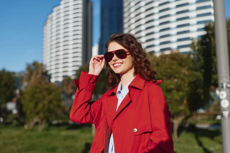 Young woman with curly hair wearing a red coat and sunglasses smiles while standing outdoors near modern buildings on a sunny day.