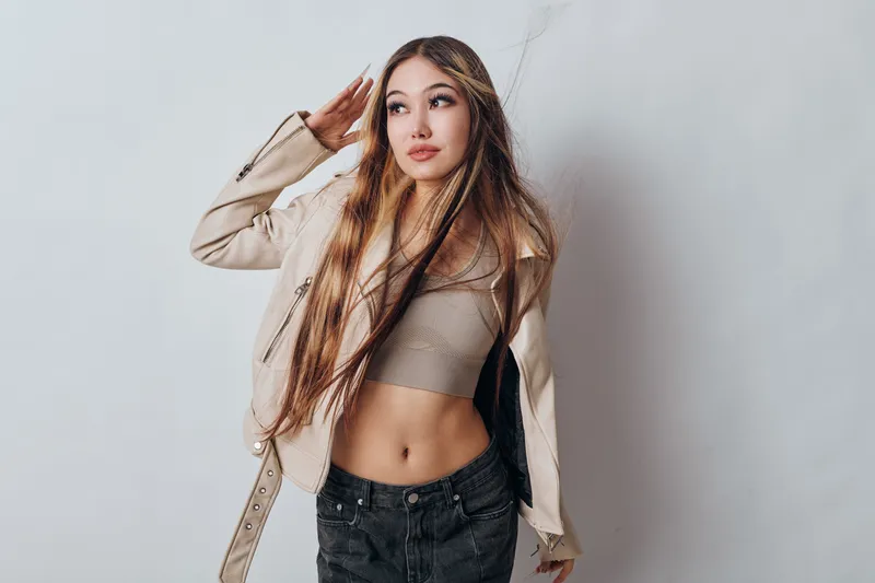 Young woman with long hair wearing casual crop top and light jacket posing with hand near head against plain background in studio portrait.