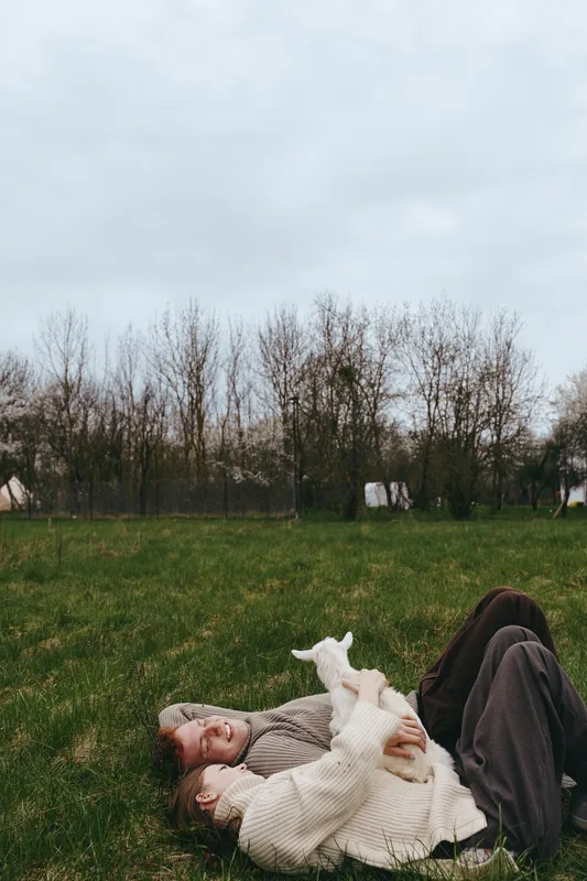 Lifestyle scene with a relaxed woman lying on grass, warmly dressed, embracing a curious goat kid in an open field, soft natural light, calm atmosphere, rustic countryside vibe