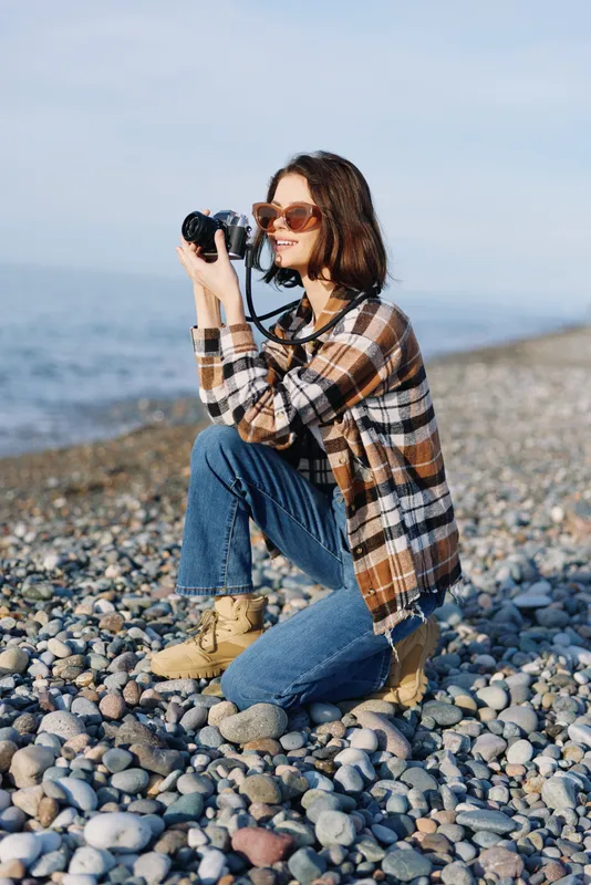 Woman photographer kneels on a pebble beach, holding a camera, wearing a plaid shirt, jeans and sunglasses, capturing seaside scenery and candid moments in bright natural light