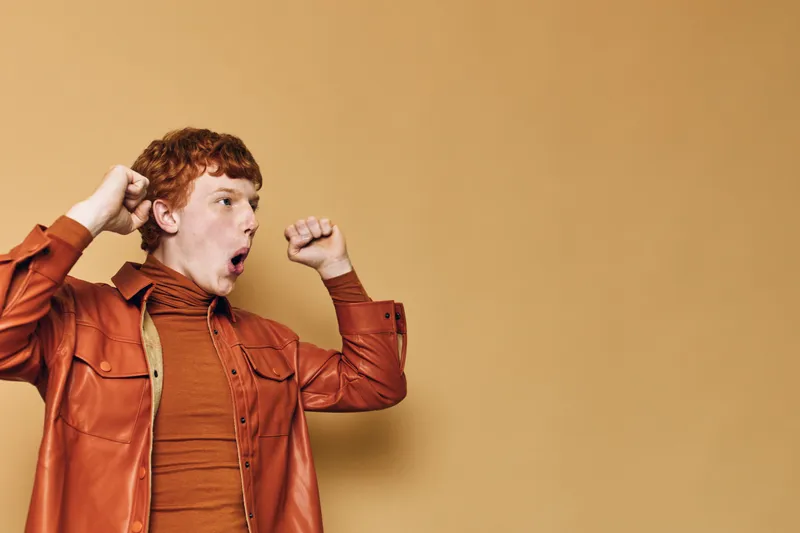 Young man in orange shirt and jacket stretching arms, yawning and waking up with tired expression in front of light brown blank background.