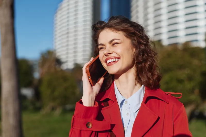 Smiling young woman with curly hair talks on a smartphone in a sunny park, wearing a red coat against an urban city backdrop.