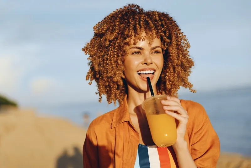 Smiling woman curly hair holds orange juice with a straw on a beach in summer outdoors, warm sunlight, seaside ocean background, casual shirt, vacation mood, joyful smile, refreshment and hydration